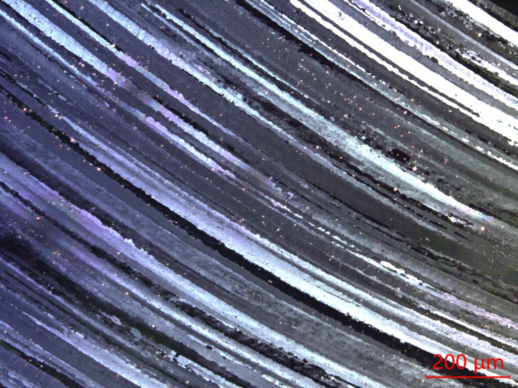 Microscopic view of layered material with wavy, iridescent lines. The scale bar indicates a dimension of 200 μm. Relevant for materials science studies.